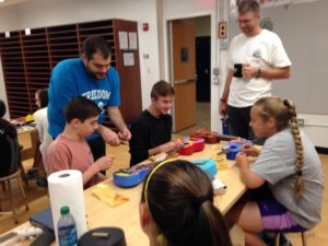 Mr. Vince Genualdi, left, helps a student learn to string his ukulele, under the watchful eye of Br. John Eustice, CSV, right